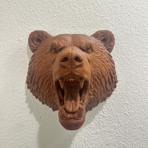 Peut inclure: Sculpture d'une tête d'ours brun avec la gueule ouverte, révélant les dents. La fourrure de l'ours est détaillée et la sculpture est fixée sur un mur blanc. L'expression de l'ours est féroce, et la sculpture est un objet décoratif.