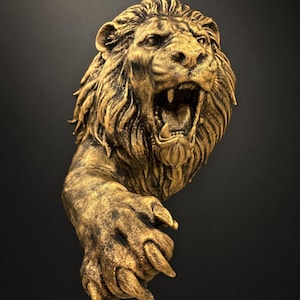 May include: A gold-coloured lion sculpture with an open mouth and detailed mane. The lion's paw is extended, showcasing intricate claw details. The sculpture is set against a dark background, highlighting the gold colour and texture.