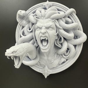 May include: A light gray, 3D-printed Medusa head sculpture. The sculpture features a woman with snakes for hair, an open mouth, and a circular frame. Two snakes are prominently displayed, one on each side of the head.
