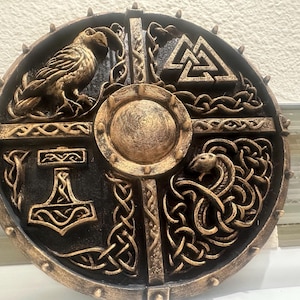 May include: A round, decorative shield with a black and gold finish. The shield features Norse symbols, including a raven, a hammer, and a serpent. The center has a circular boss. The shield is likely made of wood or a similar material.