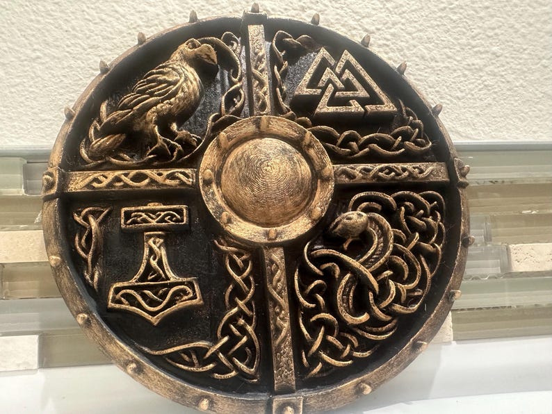 May include: A round, decorative shield with a black and gold finish. The shield features Norse symbols, including a raven, a hammer, and a serpent. The center has a circular boss. The shield is likely made of wood or a similar material.