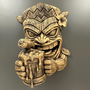 May include: A detailed, golden-brown Tiki mask sculpture with a smiling face, holding a beer stein and a cigar. The mask features carved details, a flower, and a textured finish, evoking a tropical theme.