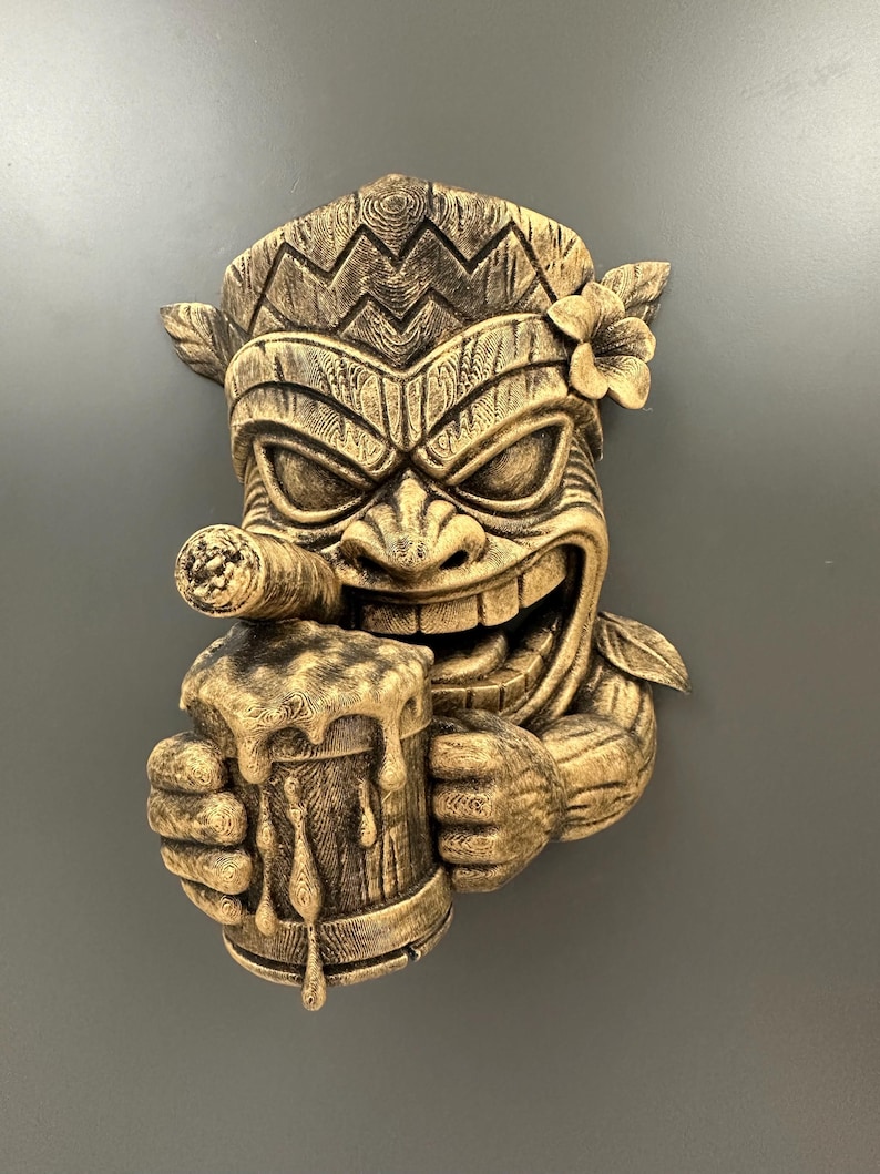May include: A detailed, golden-brown Tiki mask sculpture with a smiling face, holding a beer stein and a cigar. The mask features carved details, a flower, and a textured finish, evoking a tropical theme.