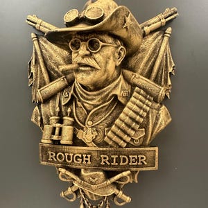 May include: A gold-colored relief sculpture of a man in a cowboy hat, goggles, and a mustache, smoking a cigar. The sculpture features crossed rifles, flags, and the text "ROUGH RIDER".