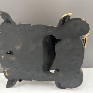 May include: Black and gold-toned decorative wall art. The piece has a carved design with a central opening and a keyhole for hanging. The edges have a gold accent.