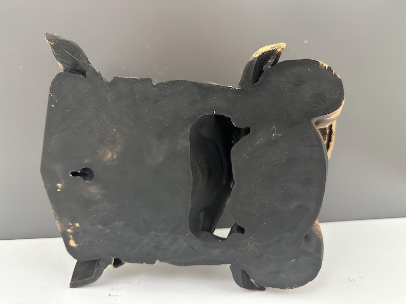 May include: Black and gold-toned decorative wall art. The piece has a carved design with a central opening and a keyhole for hanging. The edges have a gold accent.
