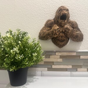 May include: A brown gorilla wall sculpture with an open mouth and detailed fur texture. The sculpture is mounted on a white wall above a decorative tile backsplash. A potted green plant sits in the foreground.