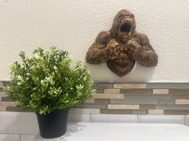 May include: A brown gorilla wall sculpture with an open mouth and detailed fur texture. The sculpture is mounted on a white wall above a decorative tile backsplash. A potted green plant sits in the foreground.