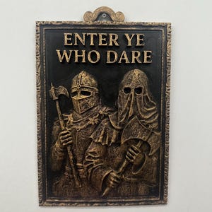 May include: A rectangular plaque with a black background and a bronze-colored frame and accents. The plaque features two figures in medieval armor holding axes, with the words "ENTER YE WHO DARE" in bold letters.