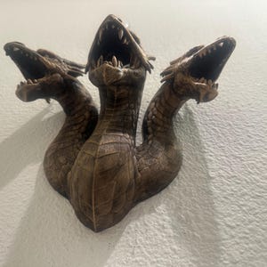 May include: A bronze-colored, three-headed dragon sculpture mounted on a wall. Each head has an open mouth with visible teeth. The sculpture has a textured surface, and the heads are positioned at different angles.
