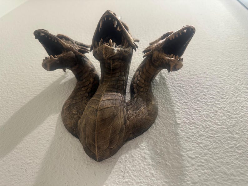 May include: A bronze-colored, three-headed dragon sculpture mounted on a wall. Each head has an open mouth with visible teeth. The sculpture has a textured surface, and the heads are positioned at different angles.