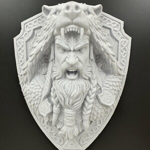 Puede incluir: Placa de pared gris impresa en 3D que representa a un guerrero vikingo con un tocado de oso y una expresión feroz. El diseño en forma de escudo incluye intrincados detalles de trenzas y nudos celtas.