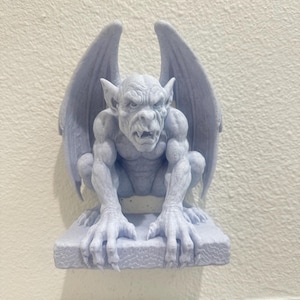 May include: A light purple gargoyle sculpture with large wings, perched on a rectangular base. The detailed statue features a fierce expression and muscular physique, perfect for gothic decor. The gargoyle is made of a solid material.