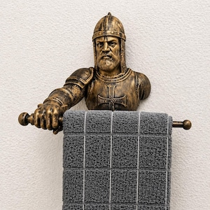 Puede incluir: Un toallero con busto de caballero de color bronce con una toalla gris. El caballero lleva un casco y una armadura, sosteniendo una barra de toalla. La toalla es gris con un patrón de cuadrícula y rayas blancas.