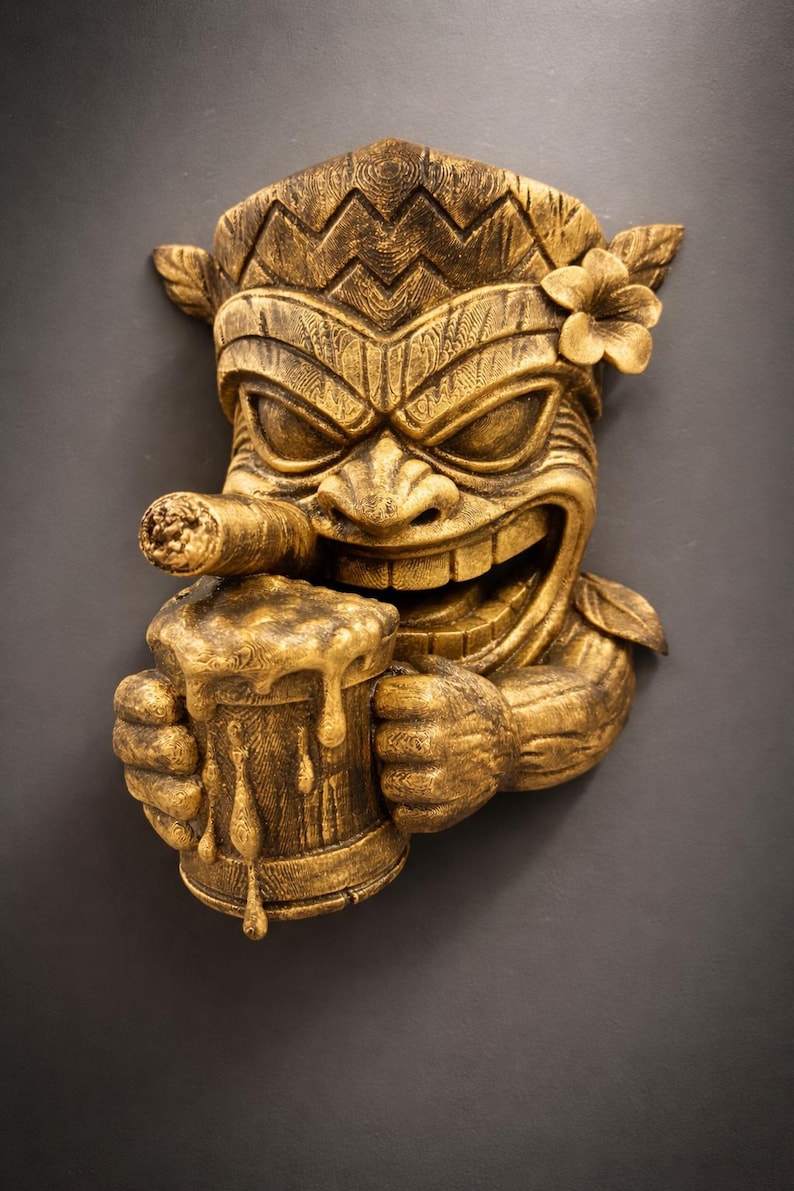 May include: A golden-colored Tiki mask sculpture with a smiling face, holding a beer mug and a cigar. The mask has a flower on its head and is carved with intricate details, perfect for tropical decor.