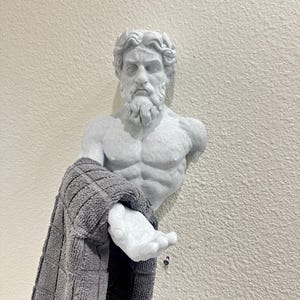 May include: A white, sculpted bust of a bearded man, possibly a Greek god, with a gray towel draped over his arm. The bust is mounted on a textured, off-white wall. The towel has a checkered pattern.