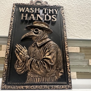 May include: Black and gold framed wall plaque with the words "WASH THY HANDS" above a relief of a plague doctor. The doctor is wearing a hat, glasses, and a long-nosed mask, with hands clasped in prayer.