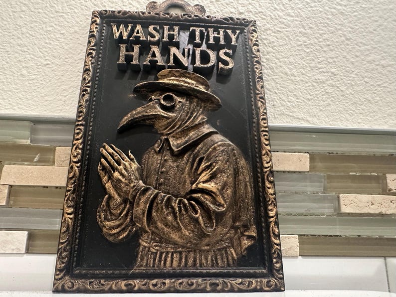 May include: Black and gold framed wall plaque with the words "WASH THY HANDS" above a relief of a plague doctor. The doctor is wearing a hat, glasses, and a long-nosed mask, with hands clasped in prayer.
