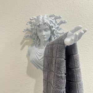 May include: A light gray Medusa head wall hook holding a gray checkered towel. The Medusa head is sculpted with detailed hair and facial features. The towel is hanging from the outstretched hand of the Medusa sculpture.