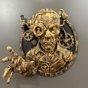 May include: A steampunk-themed wall sculpture featuring a detailed, aged face with goggles and mechanical elements. The sculpture is primarily gold and bronze, with a black circular background and gears.