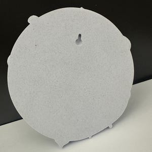 May include: A light gray, circular wall decor piece with a textured surface. The back features a keyhole for hanging. The design includes decorative elements around the edge.
