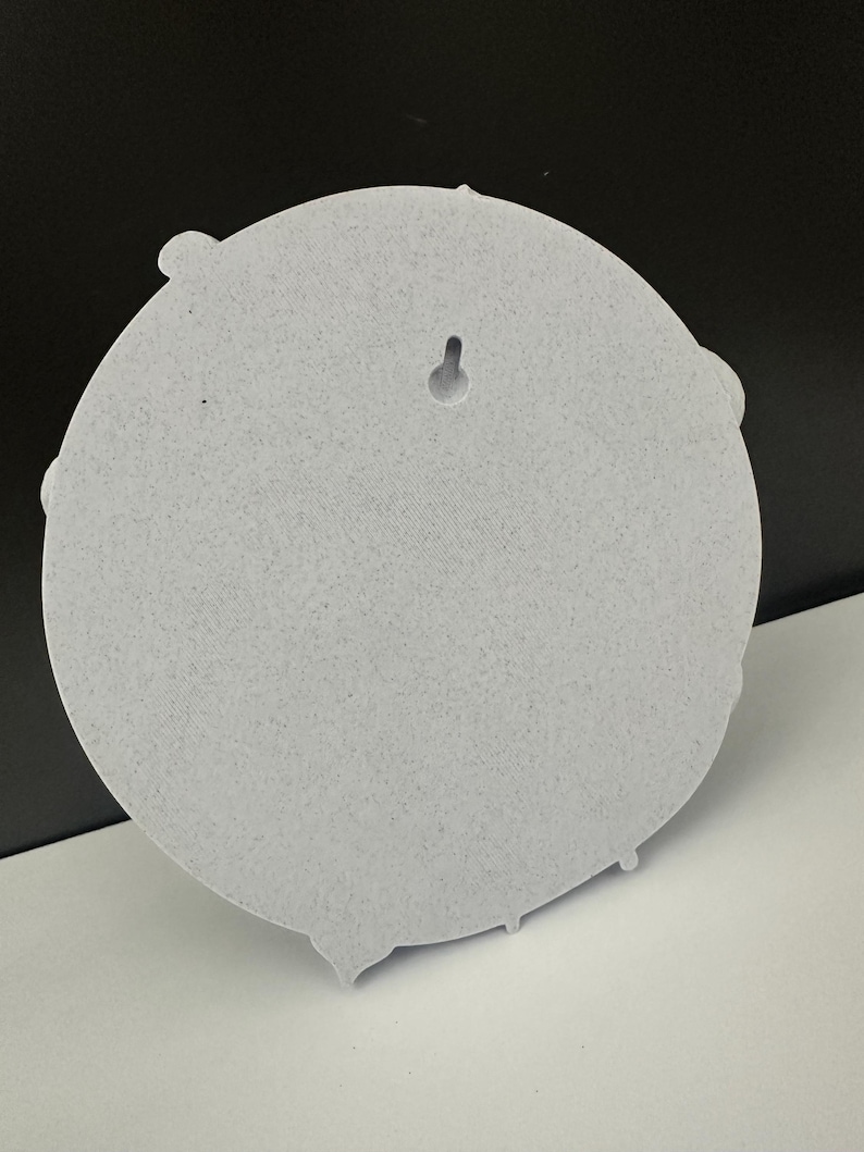 May include: A light gray, circular wall decor piece with a textured surface. The back features a keyhole for hanging. The design includes decorative elements around the edge.