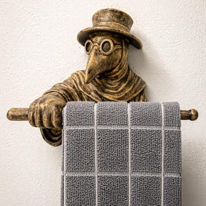 May include: A bronze-colored, decorative towel holder featuring a plague doctor figure with a long beak, round glasses, and a hat. The figure holds a gray towel with a checkered pattern. The towel holder is mounted on a white wall.