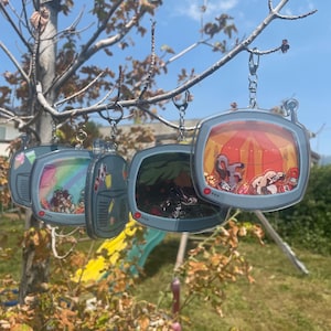 Könnte beinhalten: Mehrere Schlüsselanhänger aus Acryl in Form von Retro-Fernsehern, die jeweils eine andere farbenfrohe Szene zeigen. Die Schlüsselanhänger hängen an einem Ast vor blauem Himmel. Die Fernsehbildschirme zeigen verschiedene Bilder.