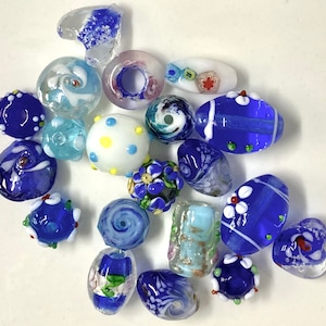 May include: A collection of colorful glass beads in various shapes and sizes. The beads feature shades of blue, white, and clear glass, with decorative swirls, dots, and floral designs. Some beads are round, oval, or heart-shaped.