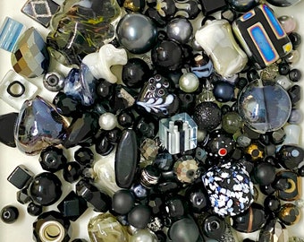 May include: A collection of black, white, and gray glass beads, some with intricate designs. The beads are a mix of shapes and sizes, including round, square, and heart-shaped.
