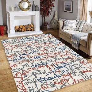 May include: A rectangular area rug with a cream background and a pattern of overlapping script in red, blue, and gray. The rug is placed on a hardwood floor in a living room setting.