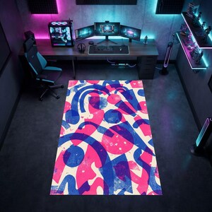 Puede incluir: Una alfombra rectangular con un patrón abstracto audaz de formas geométricas y líneas curvas superpuestas en azul y rosa intenso sobre un fondo color crema, colocada sobre un suelo oscuro en una habitación con escritorio y equipo de juego.