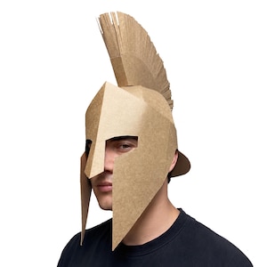 May include: A tan cardboard helmet featuring a classic Corinthian design with a prominent vertical crest. The helmet is constructed from folded brown paper material, showcasing angular geometric lines and a protective nose guard.