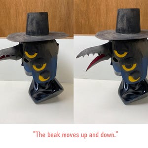 May include: A dark blue and black sculpture of a creature with a beak and hat. The beak is gray with red accents. The sculpture has three yellow eyes and the text "The beak moves up and down."