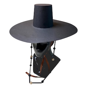 Puede incluir: Un sombrero negro con ala ancha y plana y una copa cilíndrica alta. El sombrero presenta una correa decorativa para la barbilla hecha de cordón negro, adornada con pequeñas cuentas redondas de color claro y cintas marrones que cuelgan.