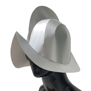 May include: A silver-colored hat with a high, rounded crown and a wide, asymmetrical brim that curves upward on one side. The hat features a smooth, metallic finish and is displayed on a black mannequin head against a plain white background.