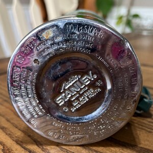 Puede incluir: Primer plano de la base de un termo Aladdin Stanley vintage. La base de metal plateado presenta texto en relieve, incluyendo "Aladdin Stanley" y "24 oz. wide mouth". El texto tambi&eacute;n incluye instrucciones de uso y el n&uacute;mero de modelo.