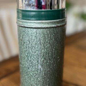 Puede incluir: Termo verde y plateado vintage. El termo tiene un cuerpo verde texturizado, una tapa plateada y una banda verde. La parte inferior tambi&eacute;n es plateada. Dise&ntilde;ado para mantener l&iacute;quidos calientes o fr&iacute;os.