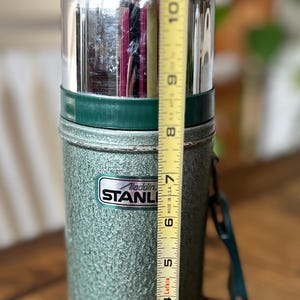 Puede incluir: Termo Aladdin Stanley verde vintage, de aproximadamente 25 cm de altura. El termo tiene un acabado texturizado, una tapa plateada y un asa verde. El logotipo de Stanley es visible en la parte delantera.