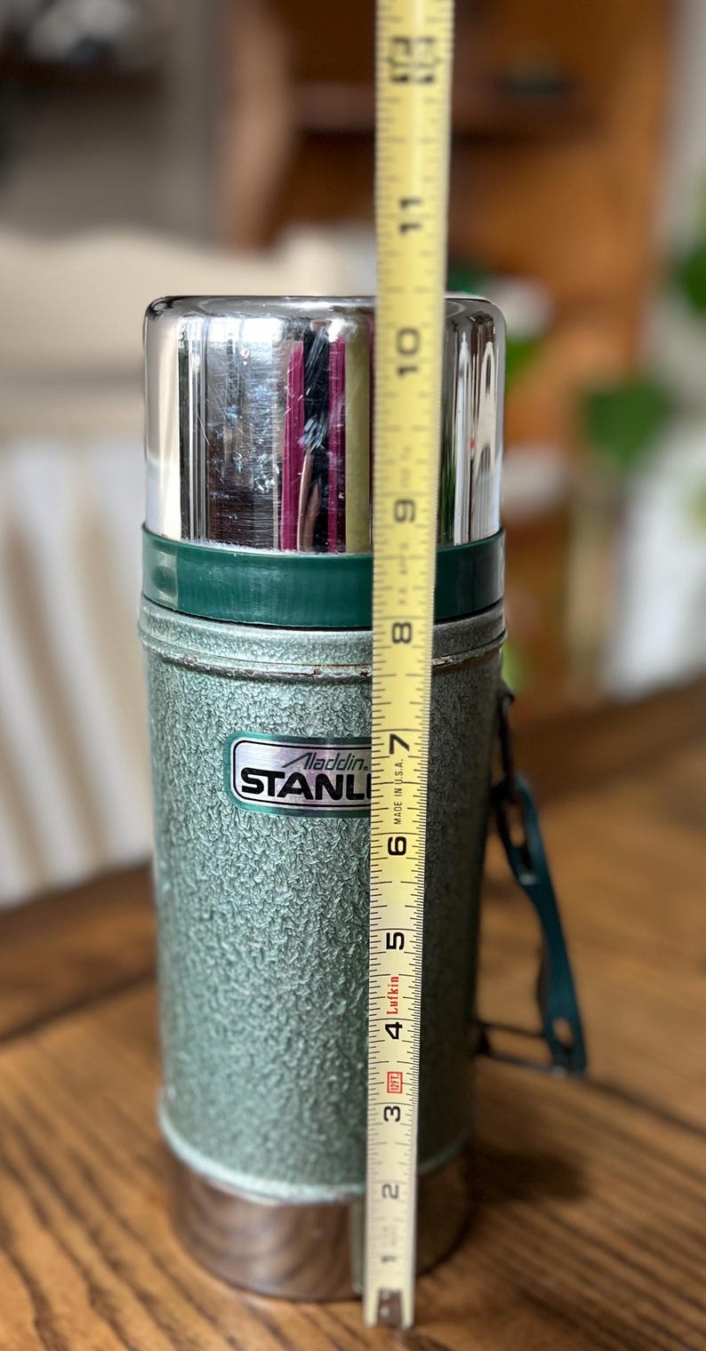 Puede incluir: Termo Aladdin Stanley verde vintage, de aproximadamente 25 cm de altura. El termo tiene un acabado texturizado, una tapa plateada y un asa verde. El logotipo de Stanley es visible en la parte delantera.