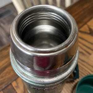 Puede incluir: Primer plano de un termo Stanley vintage. El interior de acero inoxidable es visible, rodeado por un borde de metal plateado. El exterior es de un verde descolorido con algo de desgaste. Una tapa verde es parcialmente visible.