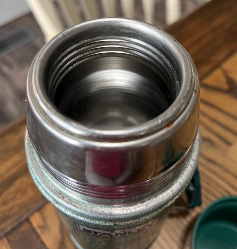 Puede incluir: Primer plano de un termo Stanley vintage. El interior de acero inoxidable es visible, rodeado por un borde de metal plateado. El exterior es de un verde descolorido con algo de desgaste. Una tapa verde es parcialmente visible.