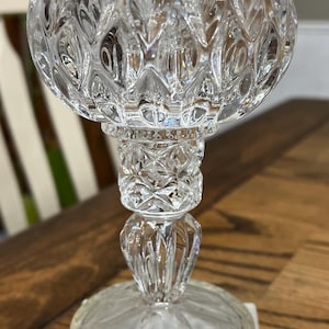 May include: A clear, ornate glass candle holder with a diamond-cut pattern. The candle holder sits atop a white marble base. The glass has a tulip shape and is designed to hold a candle. The base is rectangular.