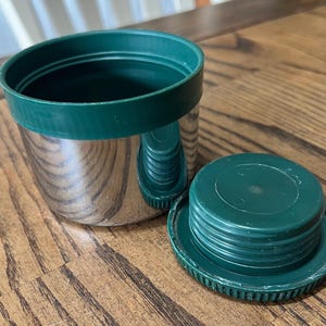 Puede incluir: Un recipiente aislado de acero inoxidable con una tapa y borde de pl&aacute;stico verde. La tapa est&aacute; a un lado, revelando el interior del recipiente. El recipiente est&aacute; sobre una superficie de madera.