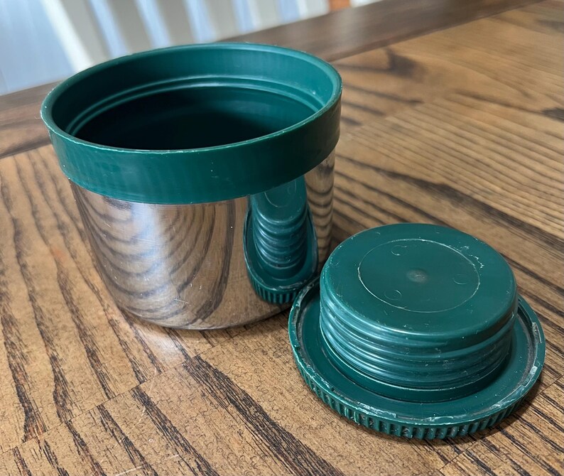 Puede incluir: Un recipiente aislado de acero inoxidable con una tapa y borde de pl&aacute;stico verde. La tapa est&aacute; a un lado, revelando el interior del recipiente. El recipiente est&aacute; sobre una superficie de madera.