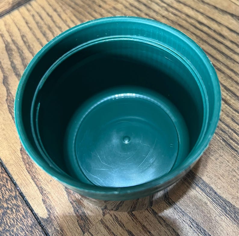 Puede incluir: Vista superior de un recipiente de pl&aacute;stico verde oscuro. El recipiente es redondo con un interior ligeramente texturizado y un exterior liso. Descansa sobre una superficie de madera.