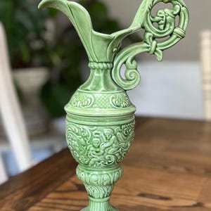 May include: A green ceramic pitcher with a decorative handle and spout. The pitcher features ornate relief carvings of cherubs and floral patterns. The base is pedestal-style, and the overall design is vintage.