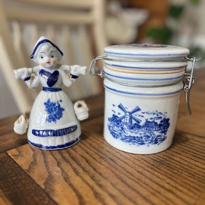 May include: A white and blue ceramic Dutch figurine and a matching jar with a metal clasp. The figurine depicts a woman in traditional attire, and the jar features a windmill scene. Both items have blue floral designs.