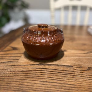 May include: A brown ceramic bean pot with a lid. The word "BEANS" is embossed on the side of the pot. The pot has two small handles and a small knob on the lid. The pot is sitting on a wooden table.