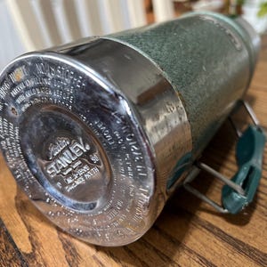 Puede incluir: Termo Stanley vintage, verde texturizado con detalles met&aacute;licos plateados. La base presenta el logo "Aladdin Stanley". El asa es verde. El termo est&aacute; sobre una superficie de madera. Dise&ntilde;ado para mantener l&iacute;quidos fr&iacute;os.